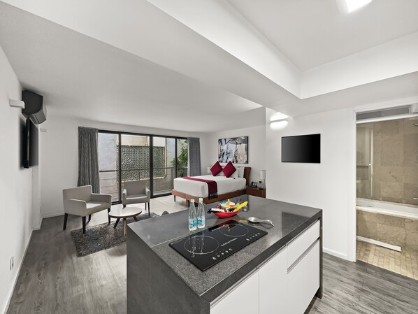 A modern hotel room with a large bed, a flat-screen TV, a dining area, and a kitchen with a built-in stove.
