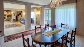 Une salle à manger spacieuse, avec une table ronde et six chaises, un lustre et une vue sur le salon, grâce à une grande fenêtre.