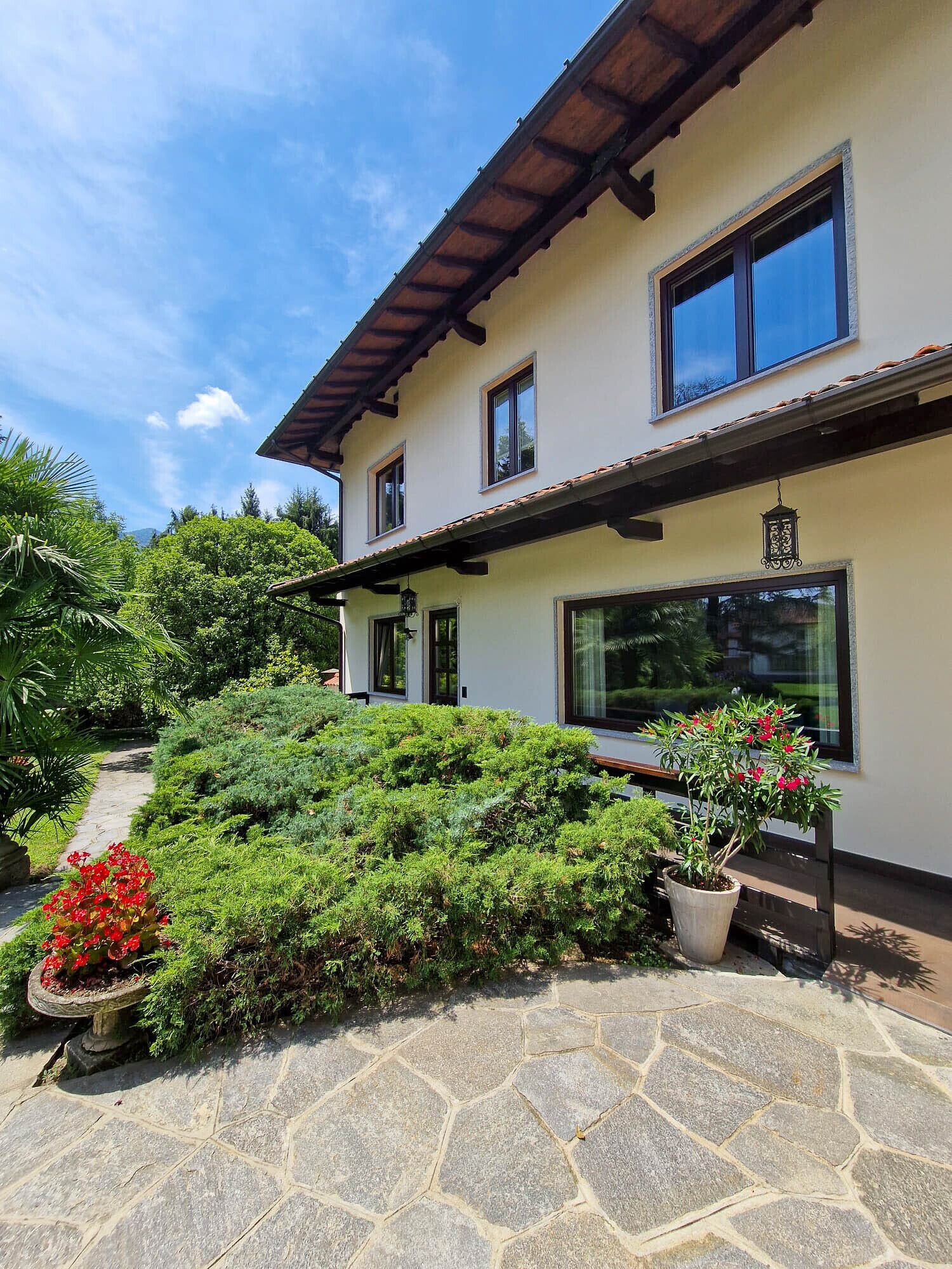 Une maison à deux étages, avec un chemin de pierre menant à l’entrée, entourée d’une végétation luxuriante et agrémentée d’un pot de fleurs aux fleurs rouges.