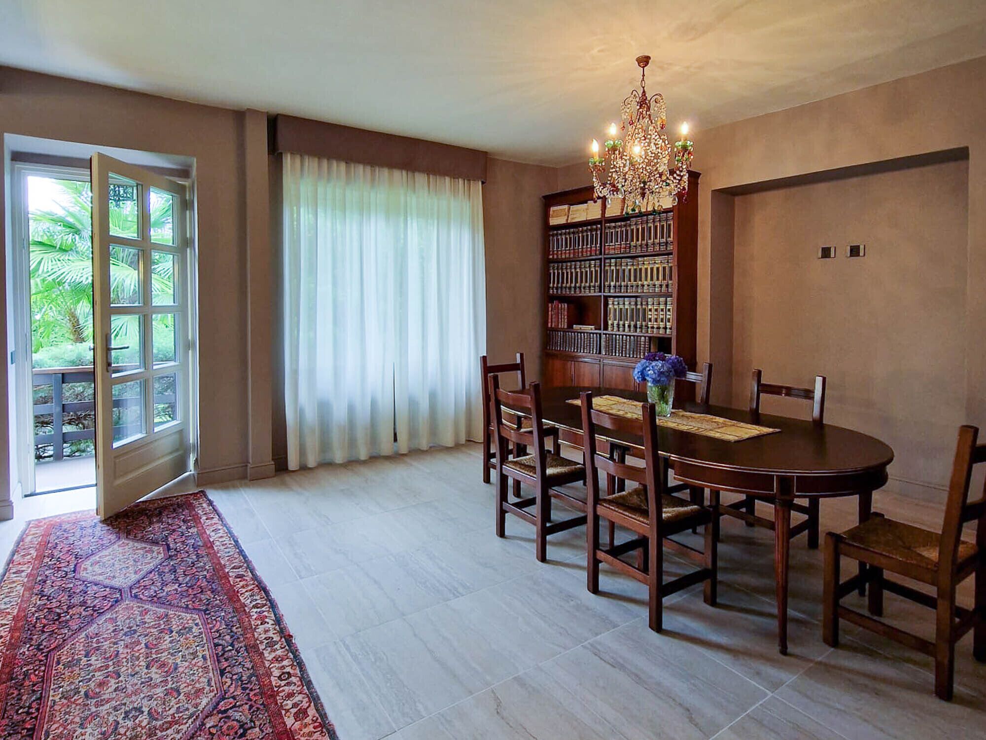A dining area with a round table, chairs, a chandelier, a bookshelf, and a view of greenery through a door.