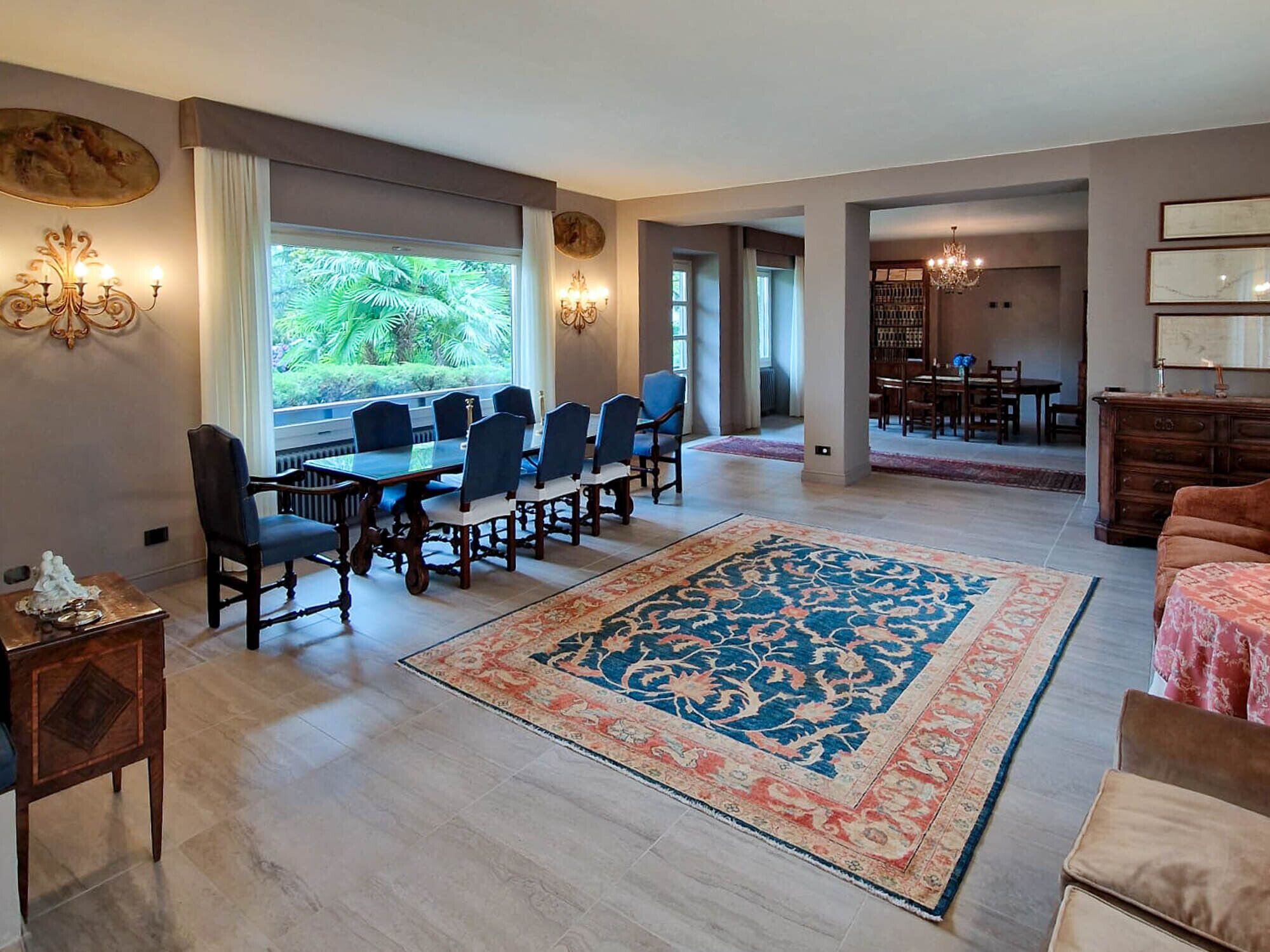 A spacious living room with a large dining table, comfortable seating, and a large window offering a view of greenery.