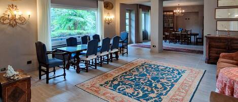 A spacious living room with a large dining table, comfortable seating, and a large window offering a view of greenery.