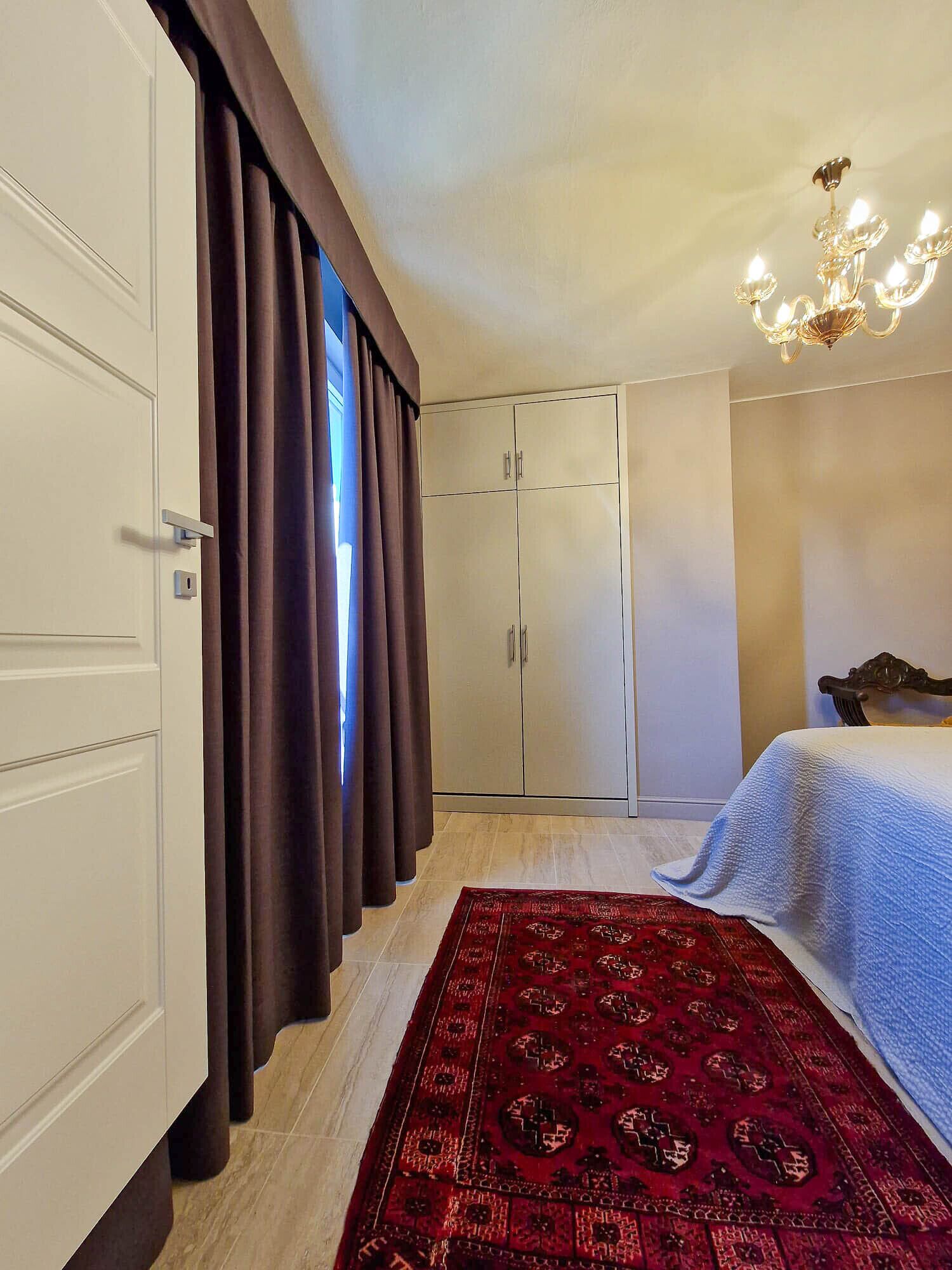 A bedroom with a bed, a wardrobe, a chandelier, and a red patterned rug.