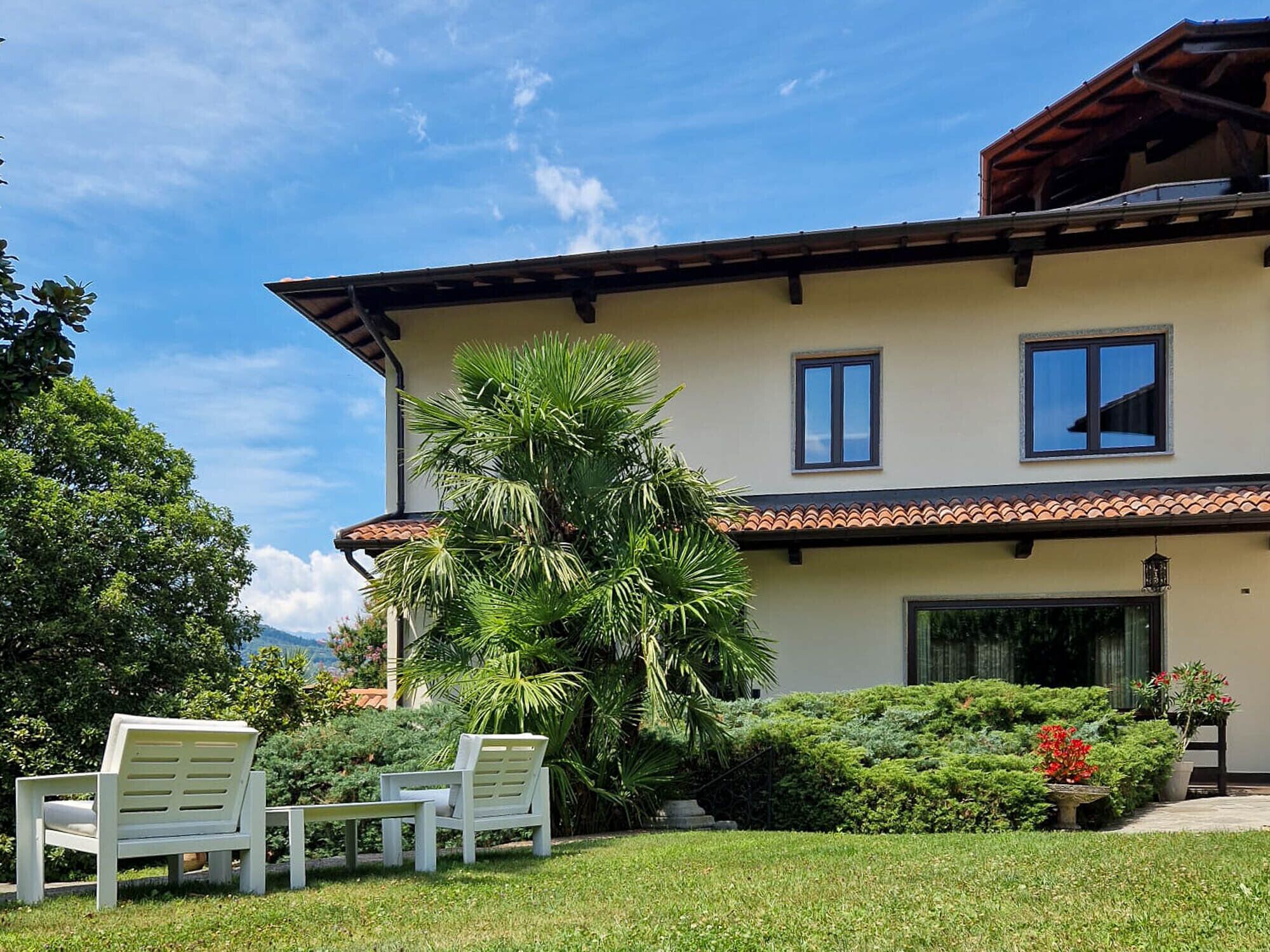 A two-story building with a tiled roof, a balcony, and a well-maintained garden with chairs and plants.