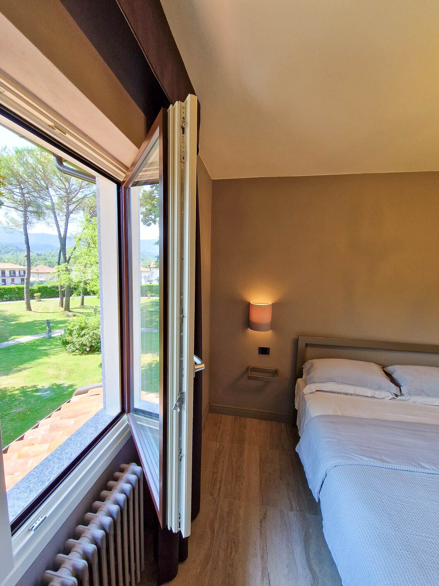 A hotel room with a large window, a bed, a bedside table with a lamp, and a radiator.