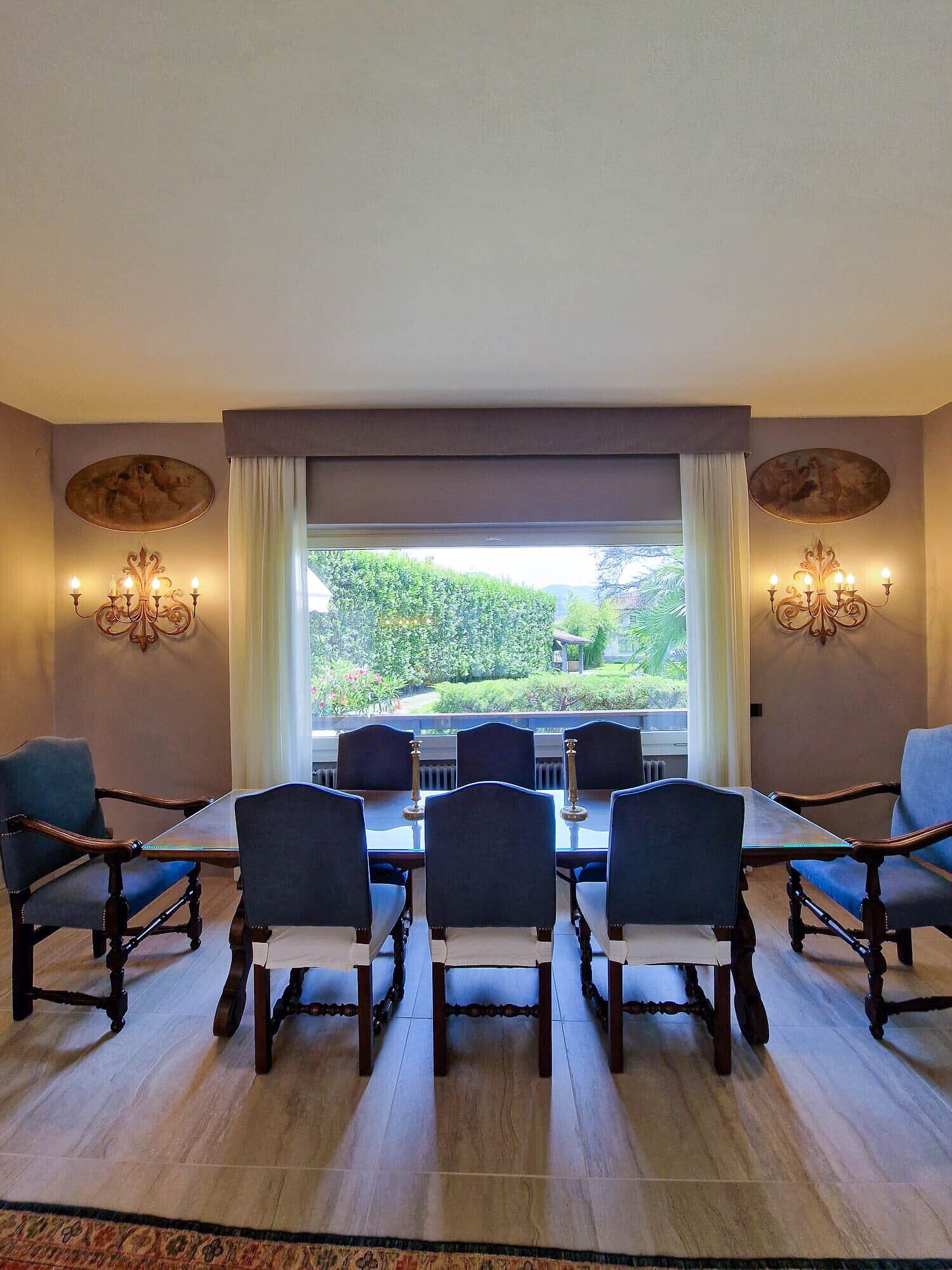 A dining room with a large table and six chairs, a chandelier, and a view of a garden through a window.