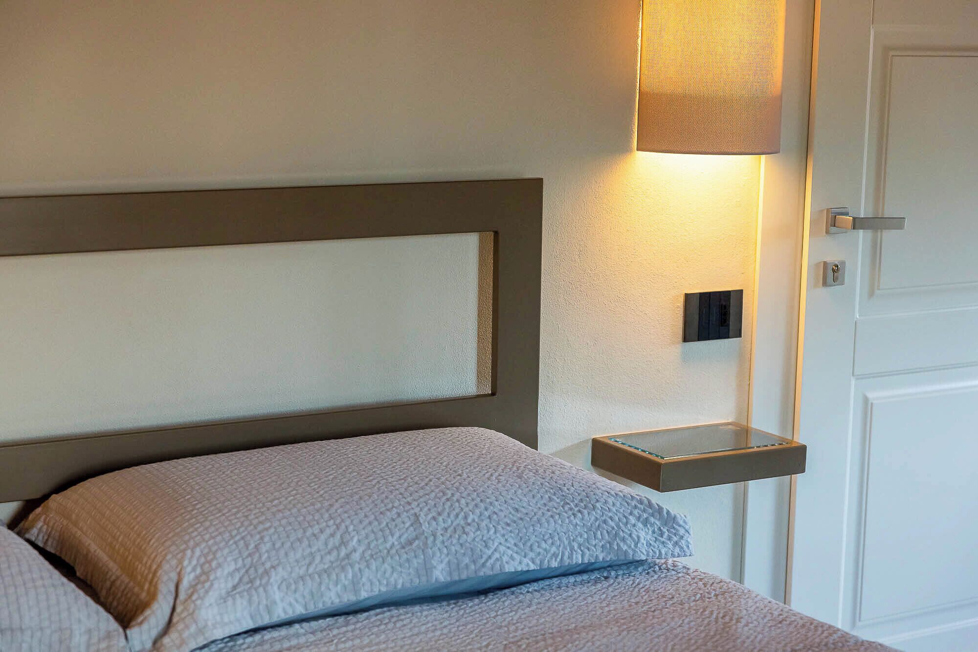 A neatly made bed with a headboard, a bedside table with a lamp, and a door in the background.