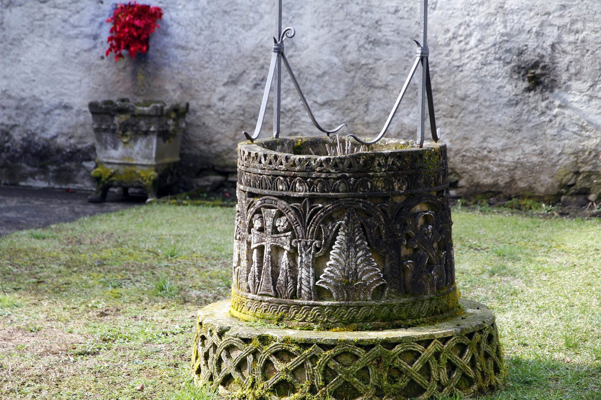 A stone structure with intricate carvings and metal hooks, possibly a historical artifact or monument, set on a grassy area with a stone wall and a red flower in the background.