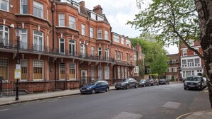 A row of red brick townhouses with a black metal fence, a tree-lined street, and parked cars.