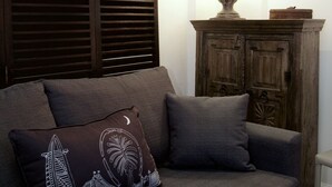 A grey sofa with a decorative pillow, a wooden cabinet with a lamp, and a window with blinds. - A Place Like Home - Inviting Flat near South Kensington (London)