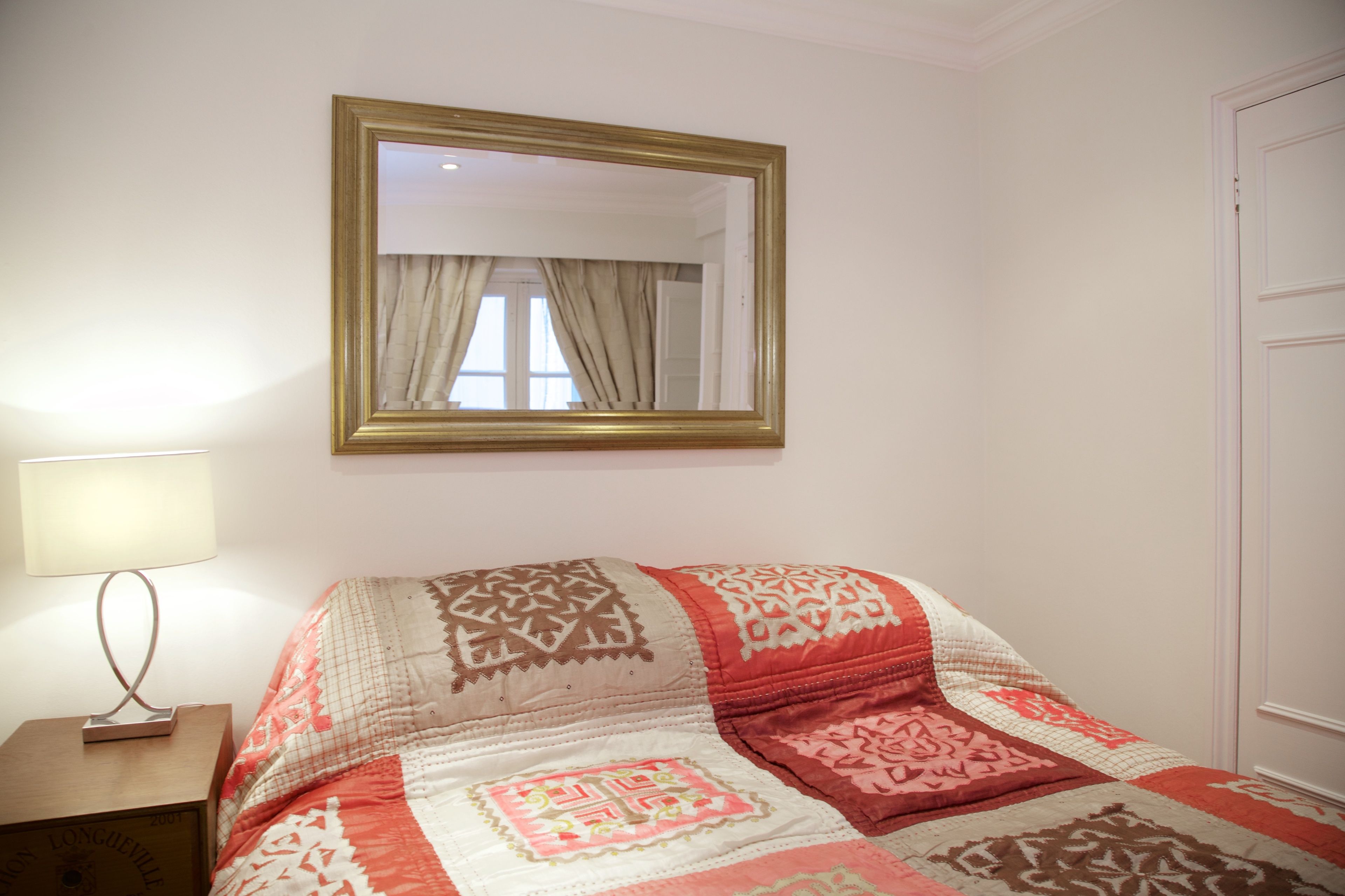 Een slaapkamer met een bed, nachtkastje, lamp, spiegel en een deur.
