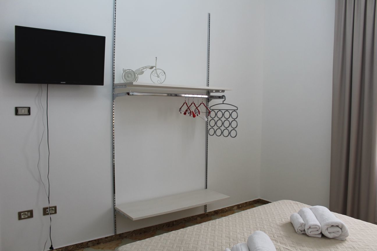 A hotel room with a bed, a TV mounted on the wall, a shelf with a bicycle and a towel rack.
