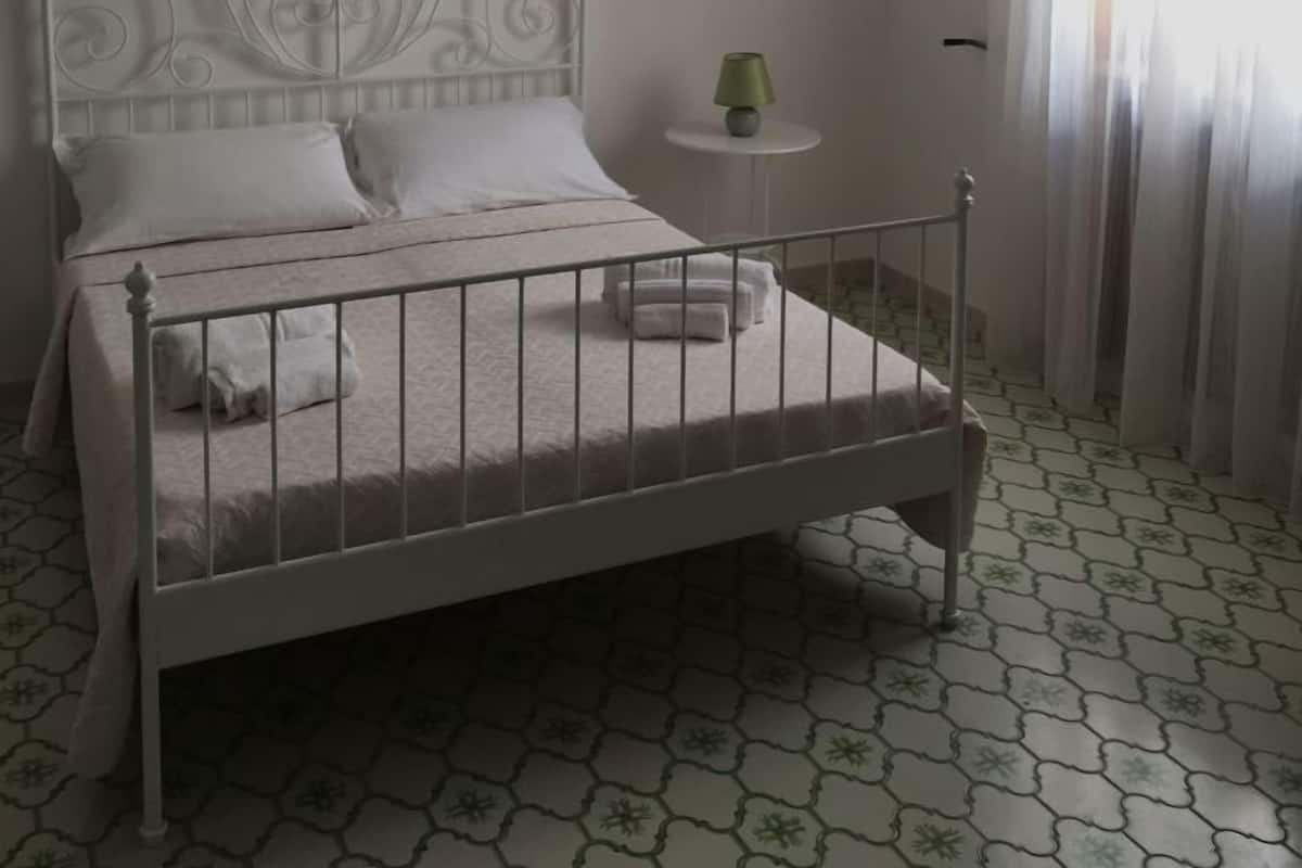 Un dormitorio con estructura de cama de metal blanco, una lámpara de techo decorativa, una mesita de noche pequeña con lámpara y una ventana con cortinas traslúcidas.