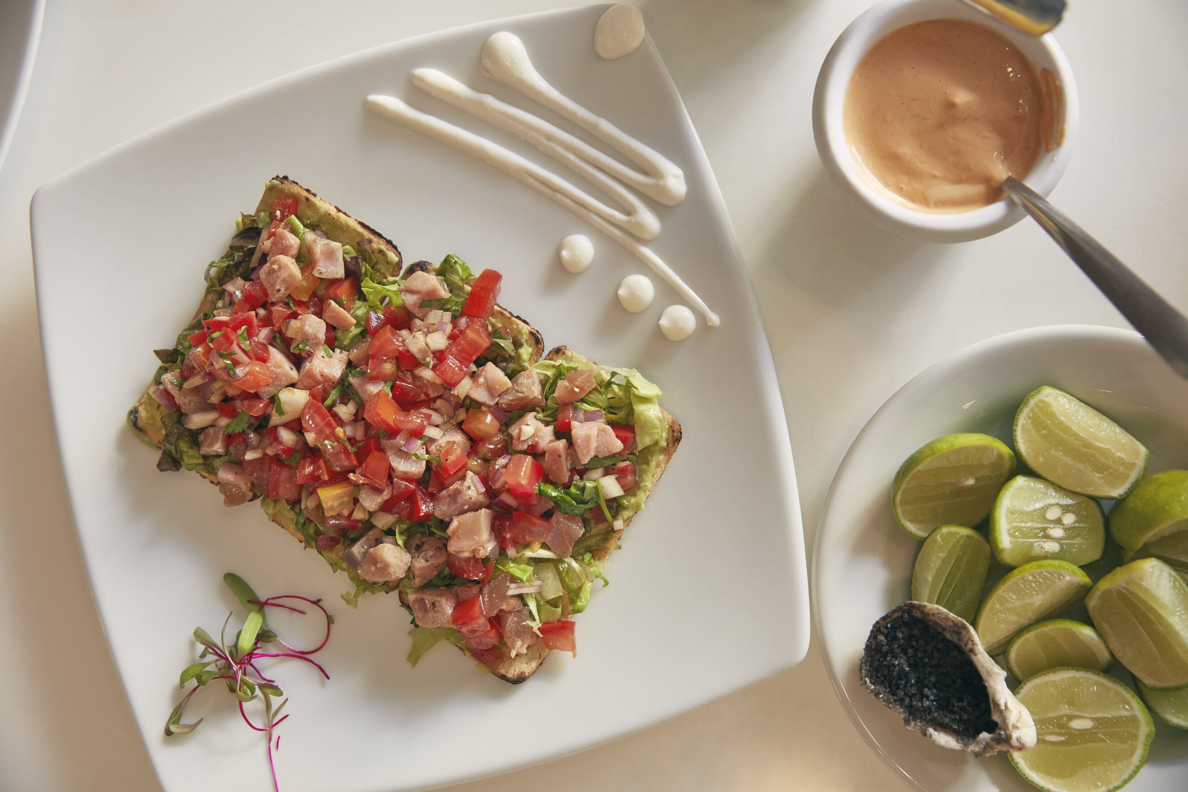 Un plato con un sándwich a la plancha cubierto de vegetales y carne picados, acompañado de gajos de lima y un recipiente pequeño con salsa.