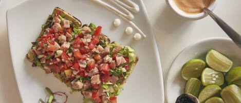 Un plato con un sándwich a la plancha cubierto de vegetales y carne picados, acompañado de gajos de lima y un recipiente pequeño con salsa.