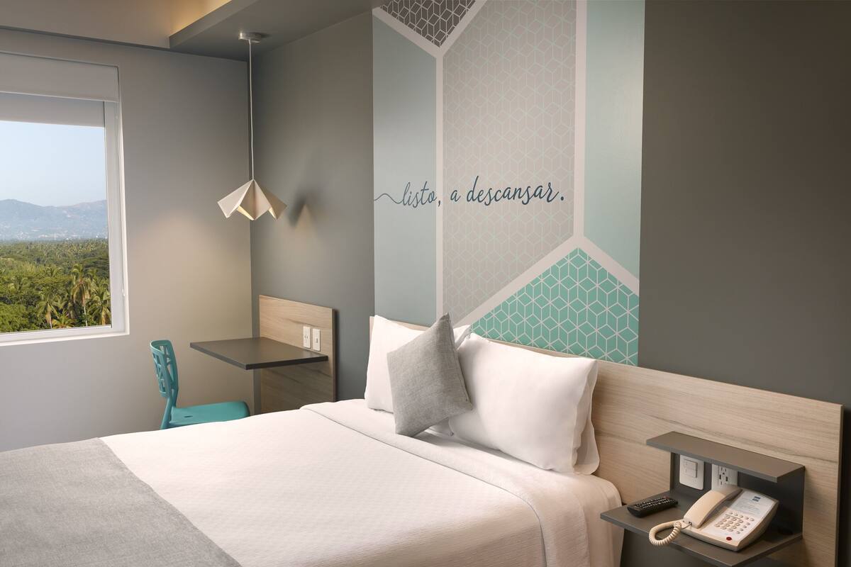 A modern hotel room with a bed, a desk, a chair, a phone, and a view of the outdoors.