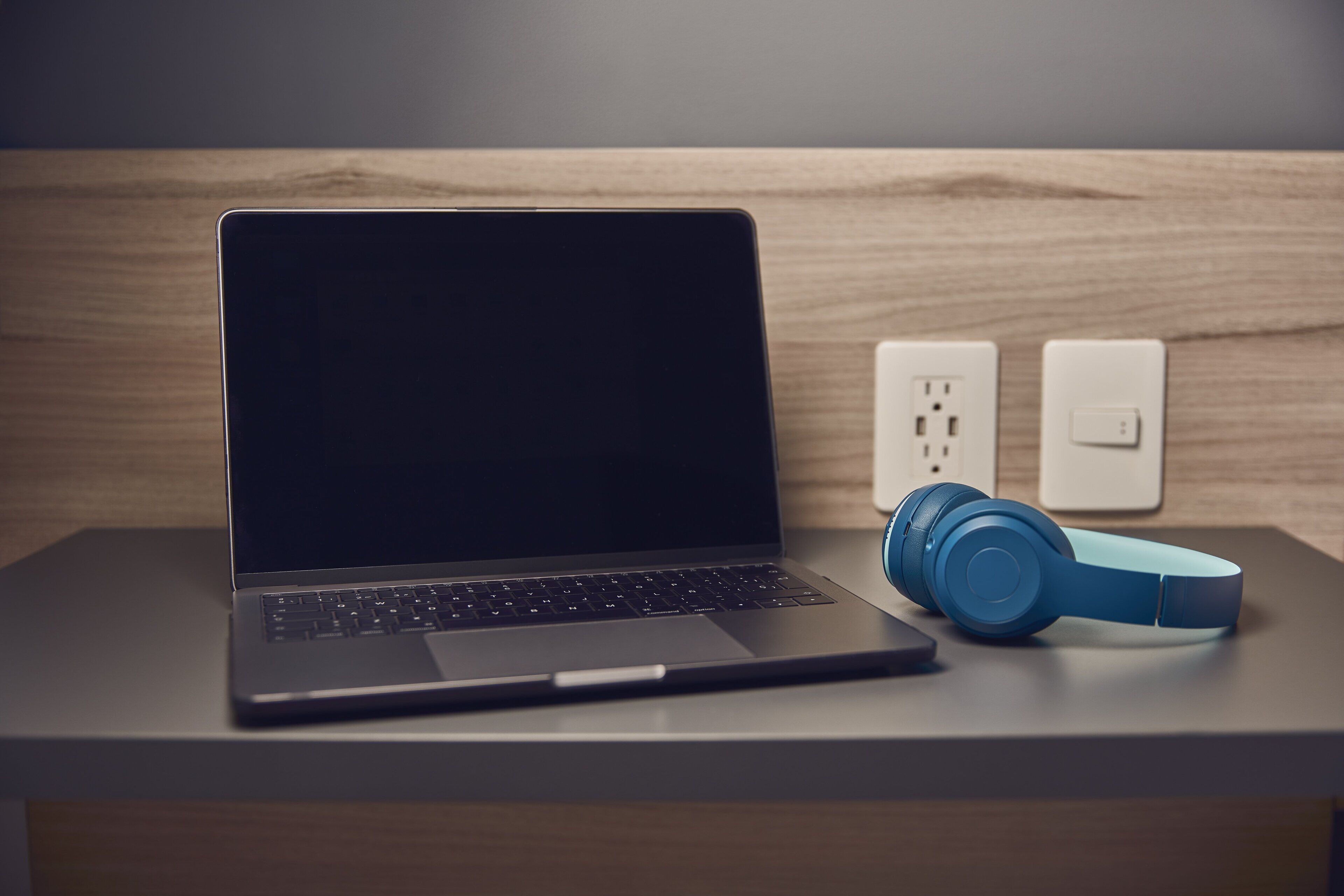 Una computadora portátil sobre un escritorio, con auriculares y un enchufe de corriente al fondo.