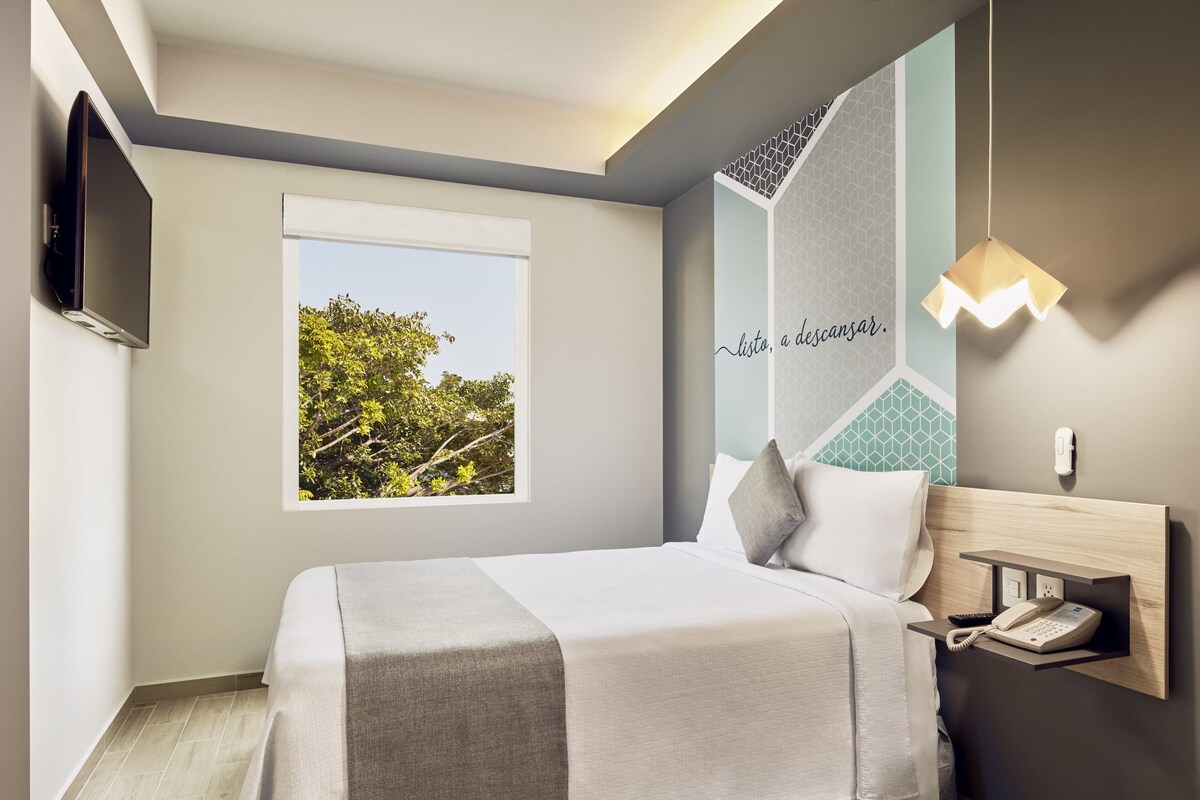 Habitación de hotel moderna con cama, televisor montado, ventana con vistas a árboles, teléfono en la pared y lámpara decorativa.