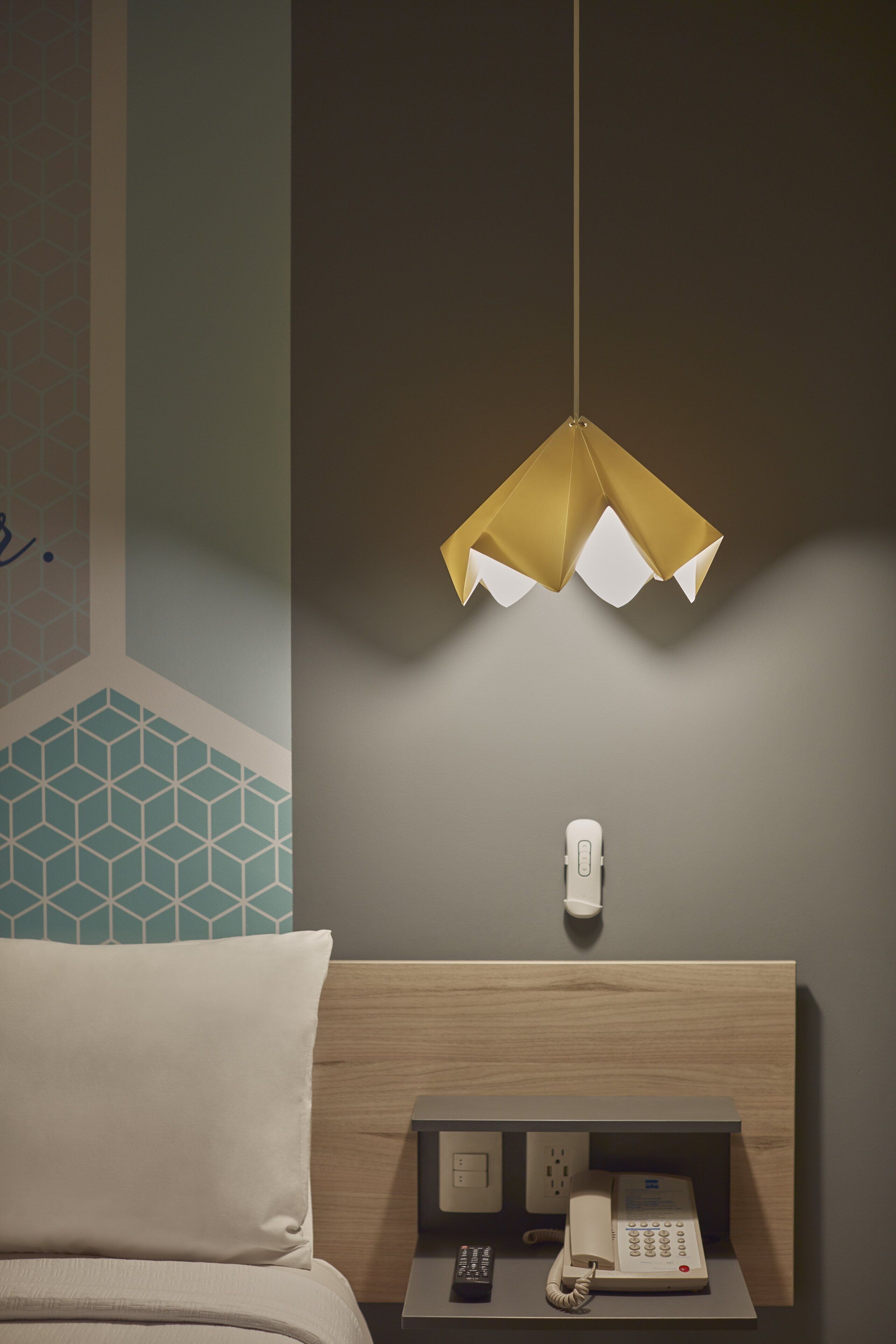A hotel room with a bed, a bedside table with a phone and remote control, a wall socket, and a hanging pendant light.