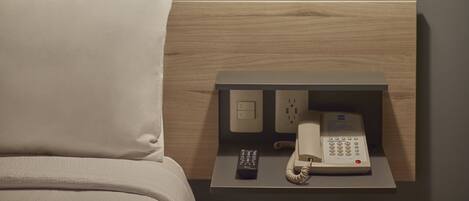 A hotel room with a bed, a bedside table with a phone and remote control, a wall socket, and a hanging pendant light.
