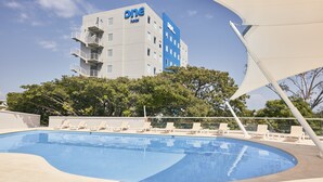 A swimming pool with lounge chairs, a building with 'one' branding, and trees in the background. - One Acapulco Diamante (Acapulco)