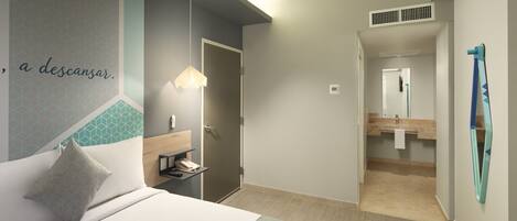 Habitación de hotel moderna con cama, mesita de noche, lámpara y vistas al baño.