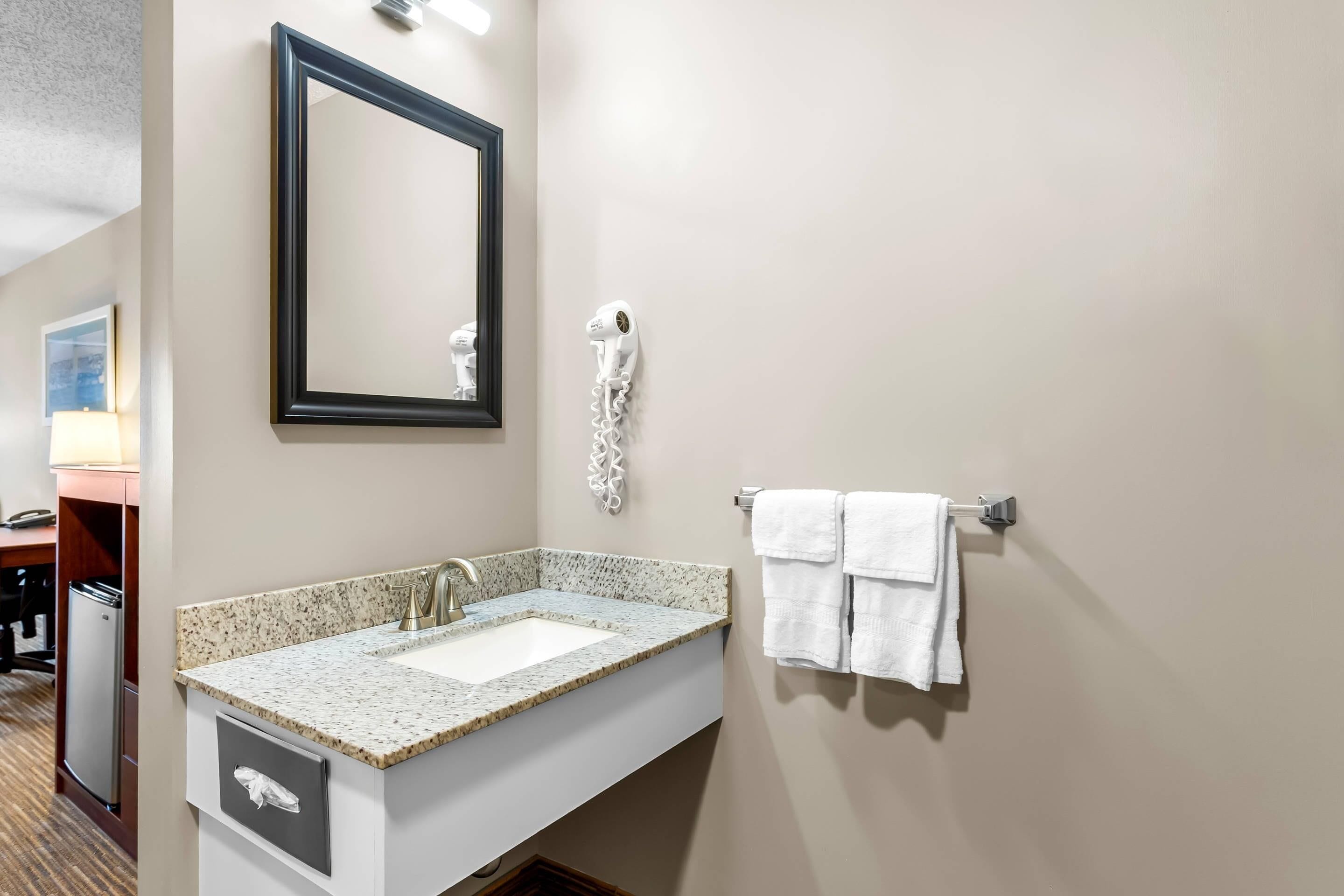 Un baño con lavamanos de granito, un espejo, un secador de pelo y un perchero para toallas.