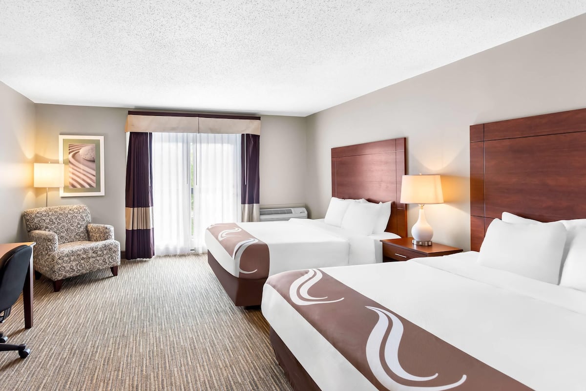 A hotel room with two beds, a desk, a chair, a large window with curtains, and a wall-mounted air conditioning unit.