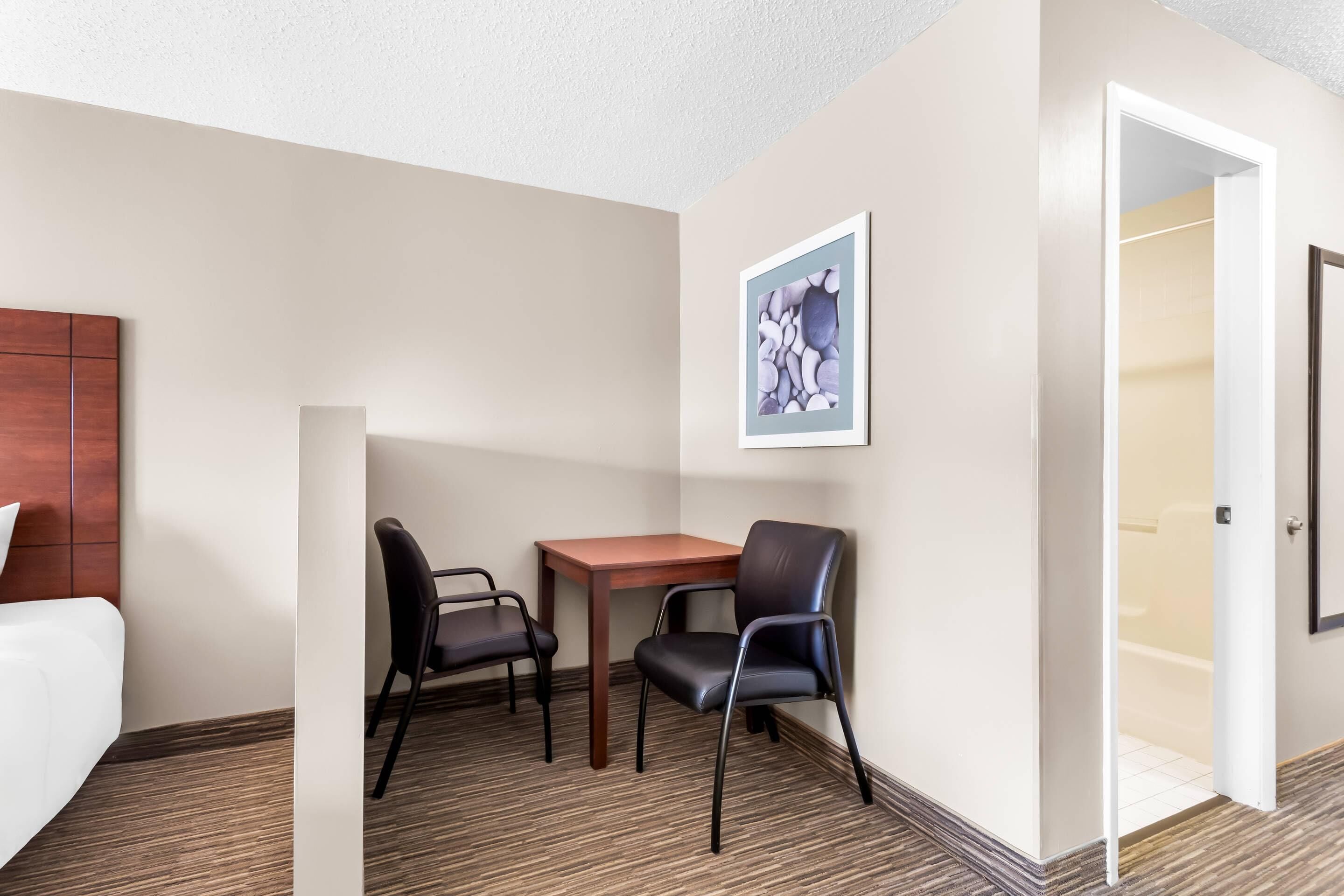 Une chambre d’hôtel avec un lit, une table en bois, deux chaises, un tableau encadré au mur et une porte donnant sur une salle de bain.