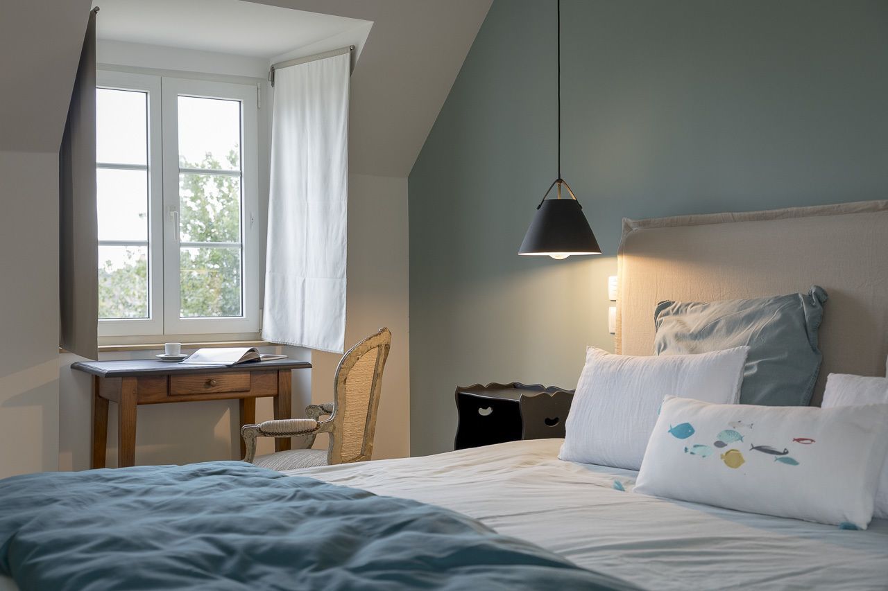 Une chambre à coucher avec un lit, un bureau et une chaise. Il y a une fenêtre avec des rideaux, une lampe suspendue et une vue sur les arbres à l’extérieur.