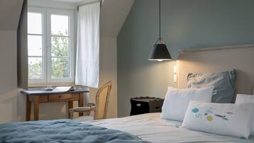 Une chambre à coucher avec un lit, un bureau et une chaise. Il y a une fenêtre avec des rideaux, une lampe suspendue et une vue sur les arbres à l’extérieur.