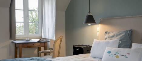 A bedroom with a bed, a desk, and a chair. There is a window with curtains, a hanging lamp, and a view of trees outside.
