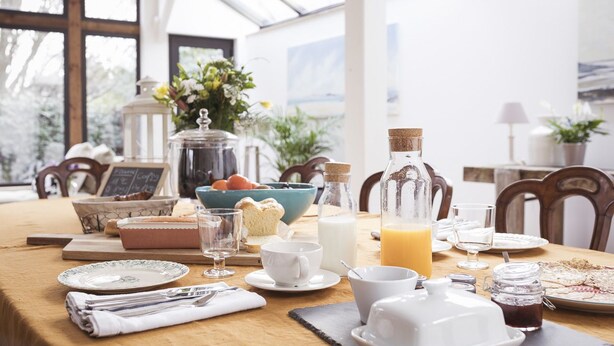 A breakfast table set with various breakfast items, including bread, eggs, milk, and juice, in a bright, naturally lit room with a view of trees outside. - Cottage Santa Teresa (Saint-Lunaire)