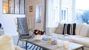 A cozy living room with a wicker chair, a white sofa with striped pillows, a coffee table with candles and a vase of flowers, and a large window offering a view of the outdoors. - Cottage Santa Teresa (Saint-Lunaire)