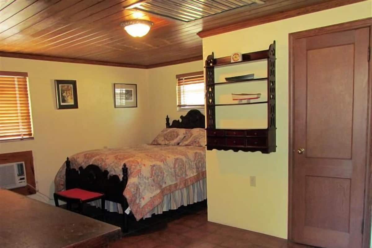 A bedroom with a bed, a wooden shelf with books, a door, and a window with blinds.