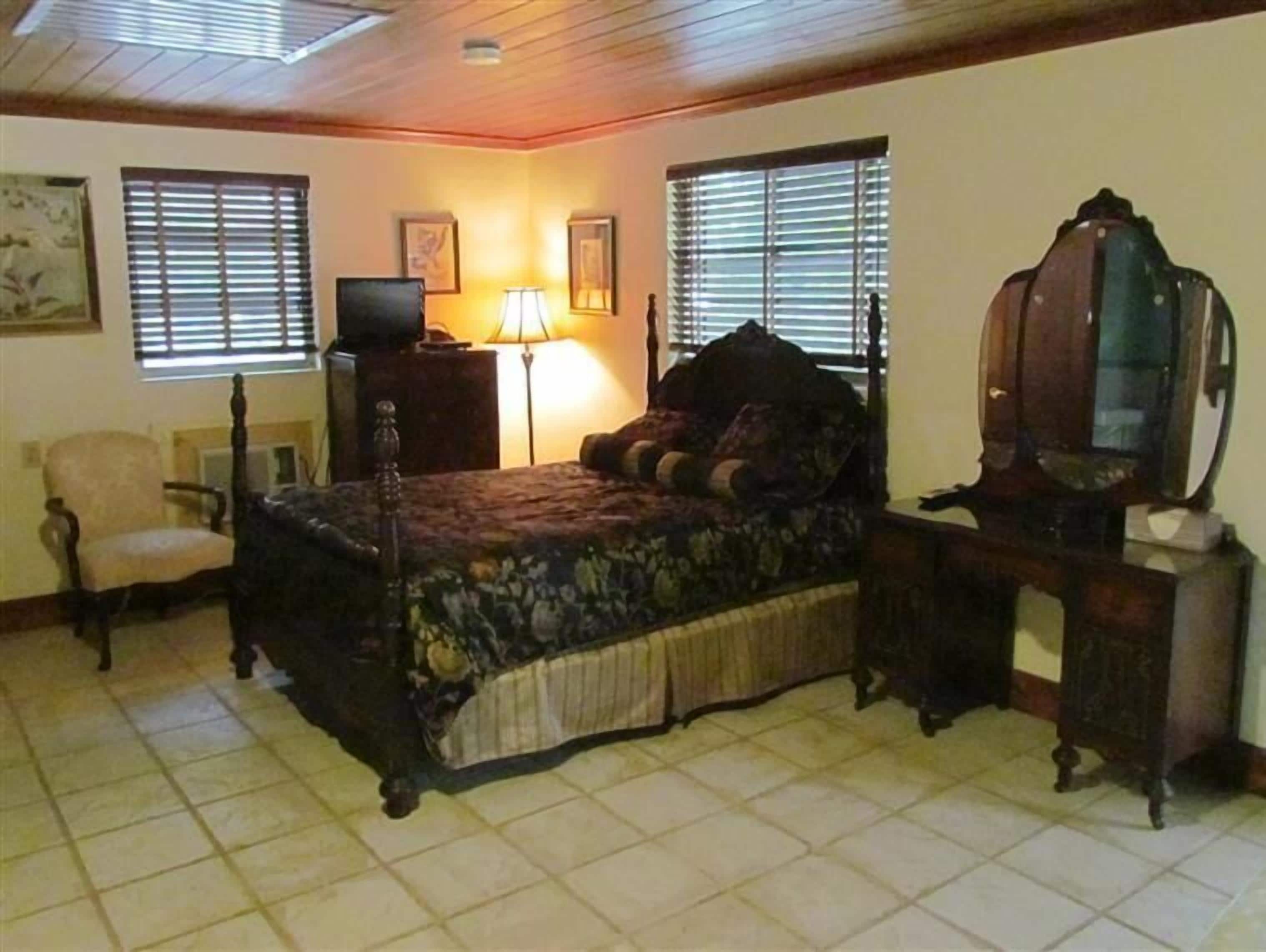 A bedroom with a bed, a chair, a desk with a television, and two windows with blinds.