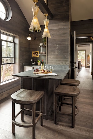 A kitchen with a central island, wooden stools, pendant lighting, and a view through a window.