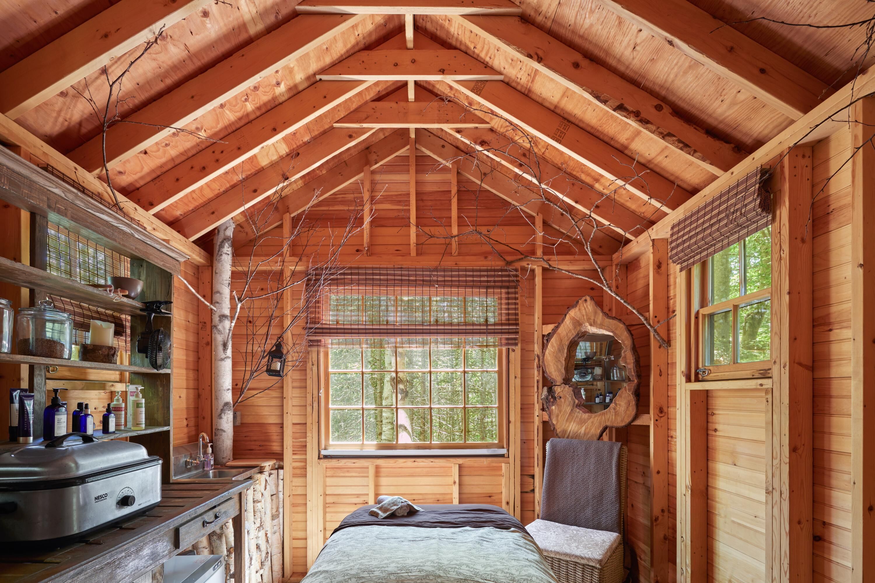 A cozy cabin interior with a wooden ceiling, a bed, a chair, and a window with a view of trees.