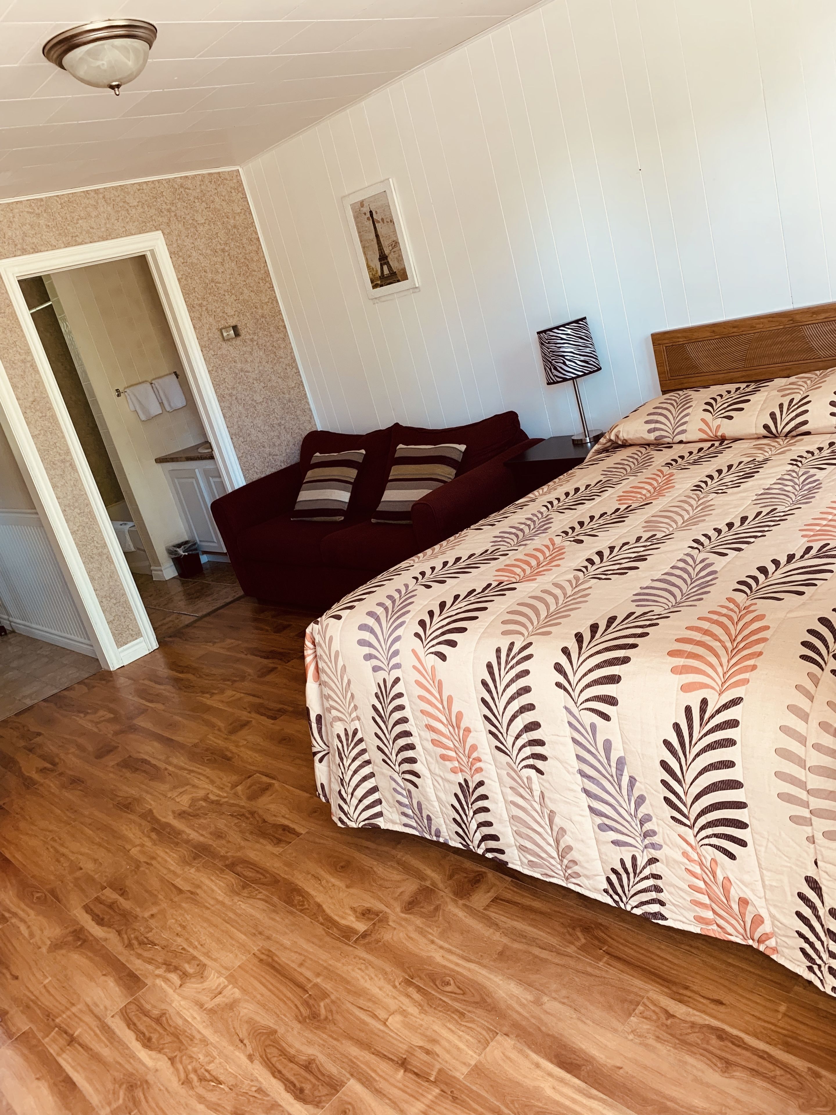 A bedroom with a wooden floor, a bed with a leaf-patterned bedspread, a dark-colored sofa with striped pillows, a framed picture of the Eiffel Tower, and a floor lamp.
