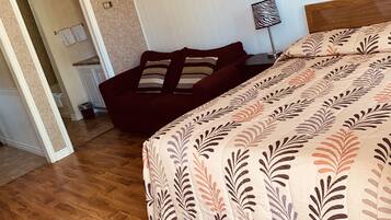 A bedroom with a wooden floor, a bed with a leaf-patterned bedspread, a dark-colored sofa with striped pillows, a framed picture of the Eiffel Tower, and a floor lamp.
