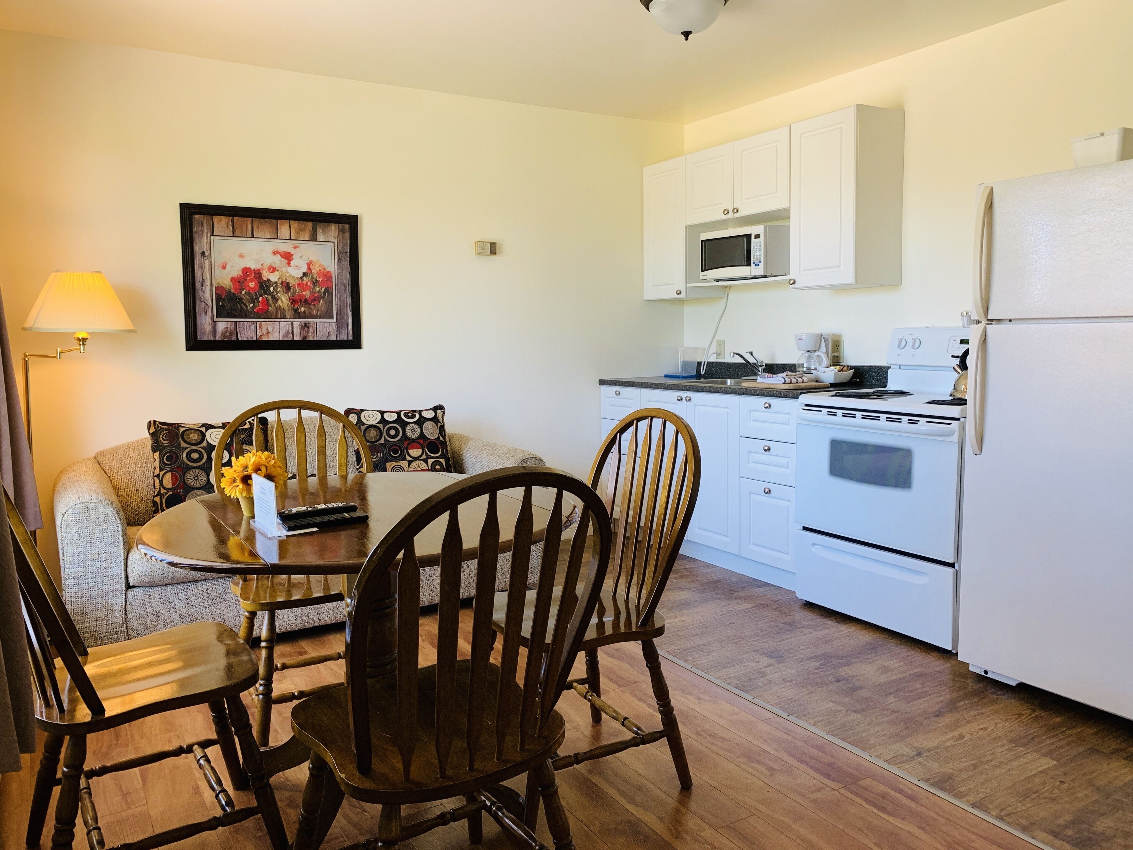 A compact kitchen with white cabinets, a microwave, a stove, and a refrigerator. There is a dining table with chairs and a lamp.