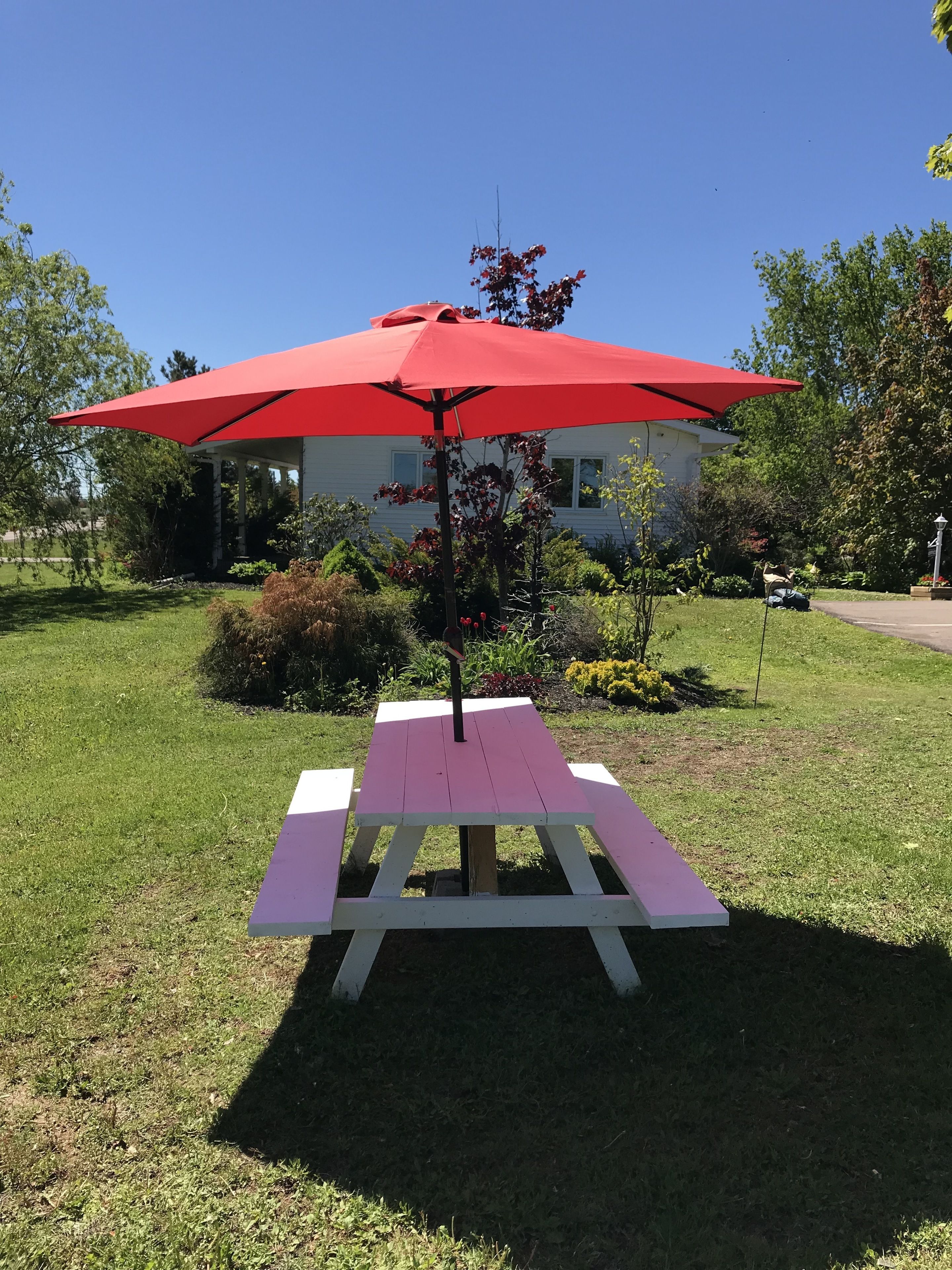 A picnic table with a red umbrella in a grassy area with trees and a house in the background.
