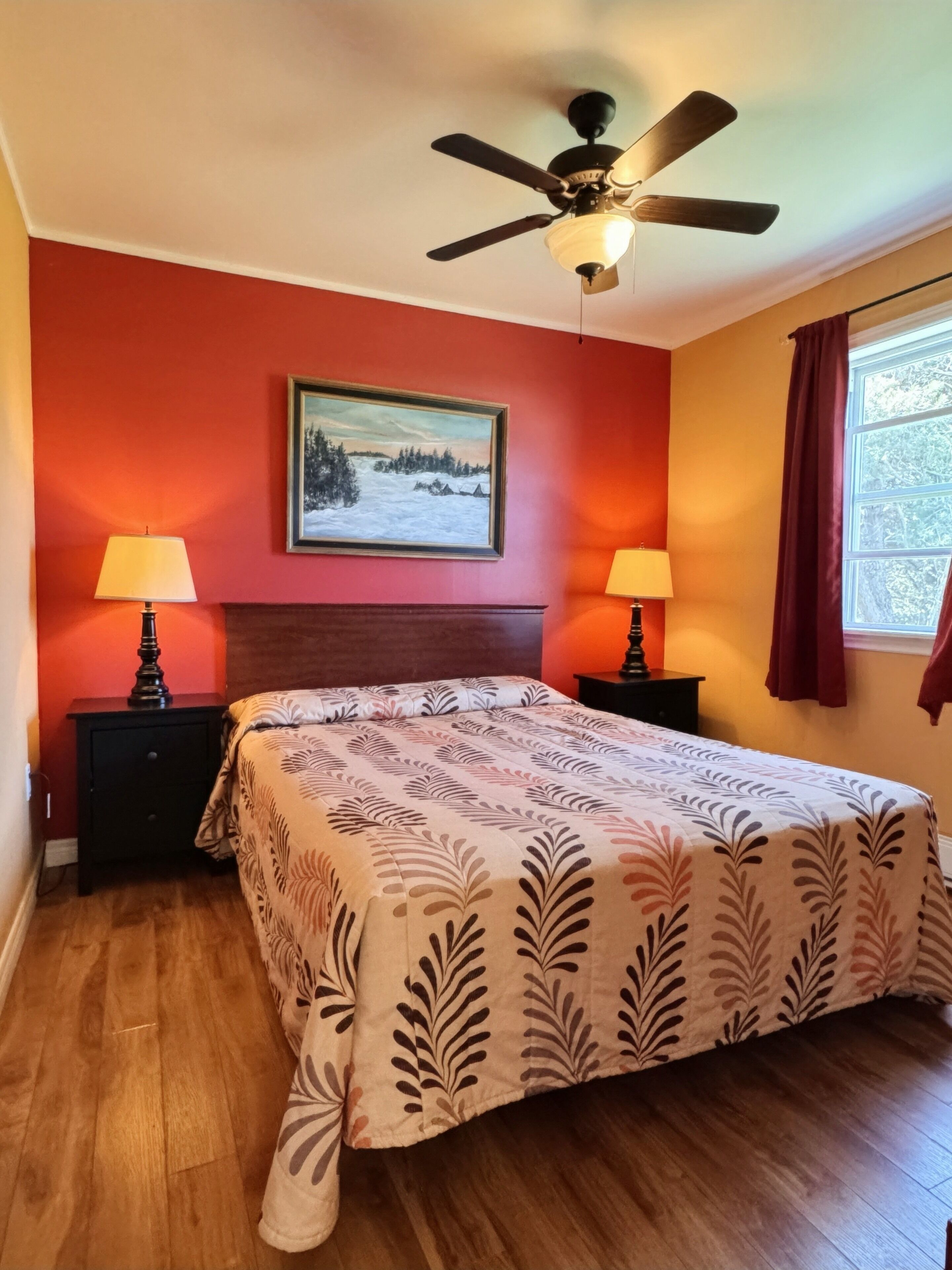 Une chambre à coucher comprenant un lit, deux tables de chevet, un ventilateur de plafond, une fenêtre avec des rideaux et un tableau accroché au mur.