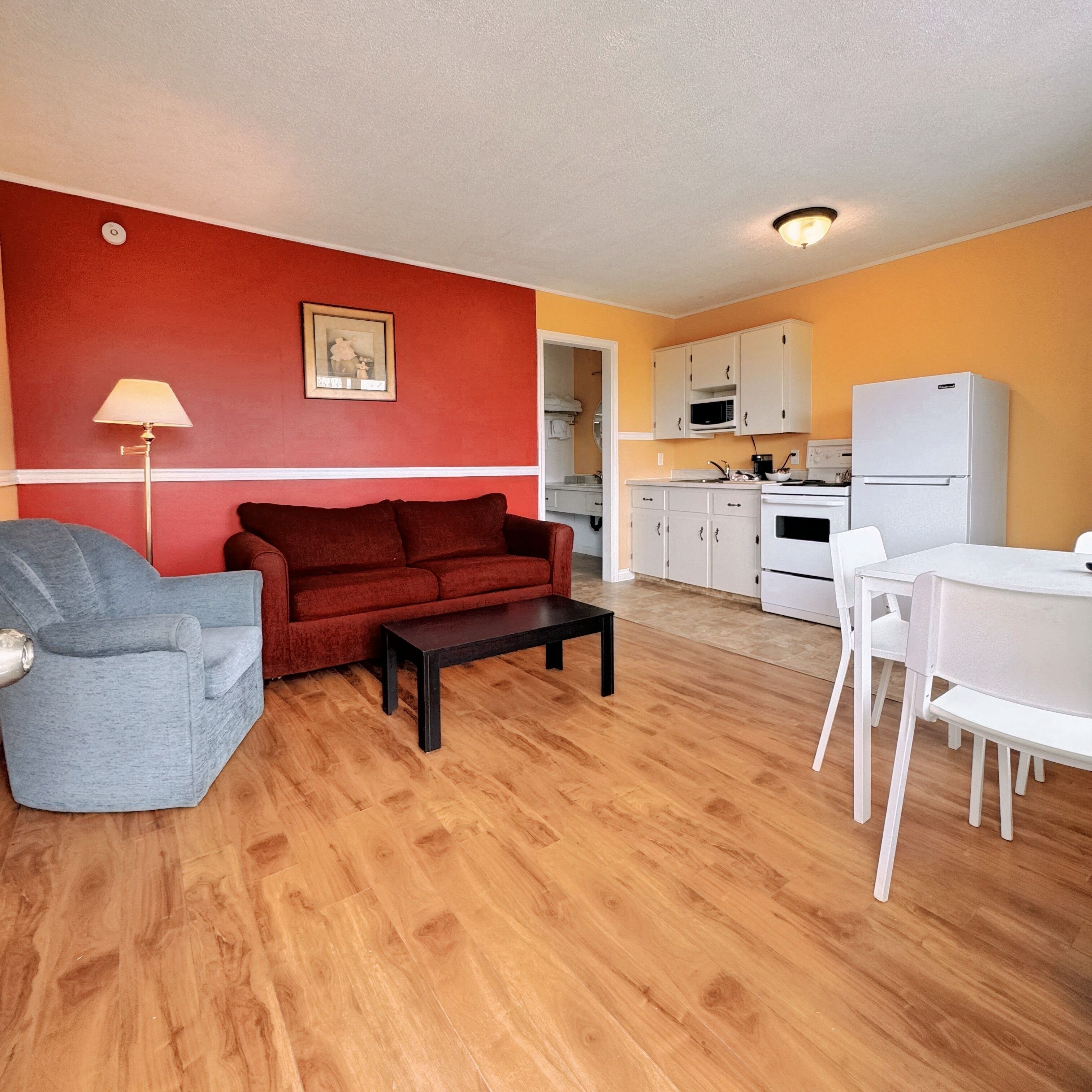 A compact living space with a red sofa, a grey armchair, a small table, a white dining table with chairs, and a kitchen area with white cabinets and appliances.