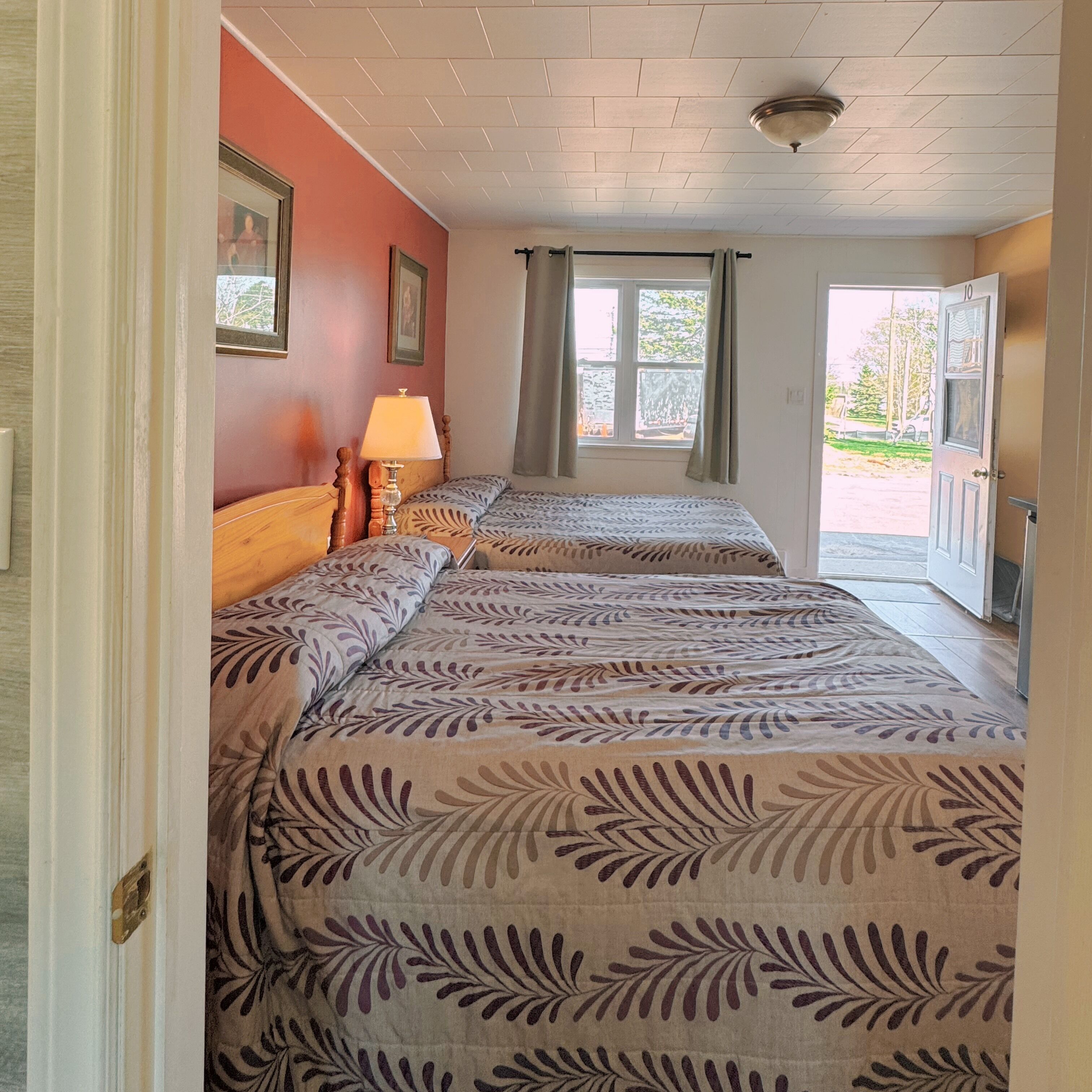 Una habitación con dos camas, una ventana con cortinas, una lámpara y una puerta que da al exterior.