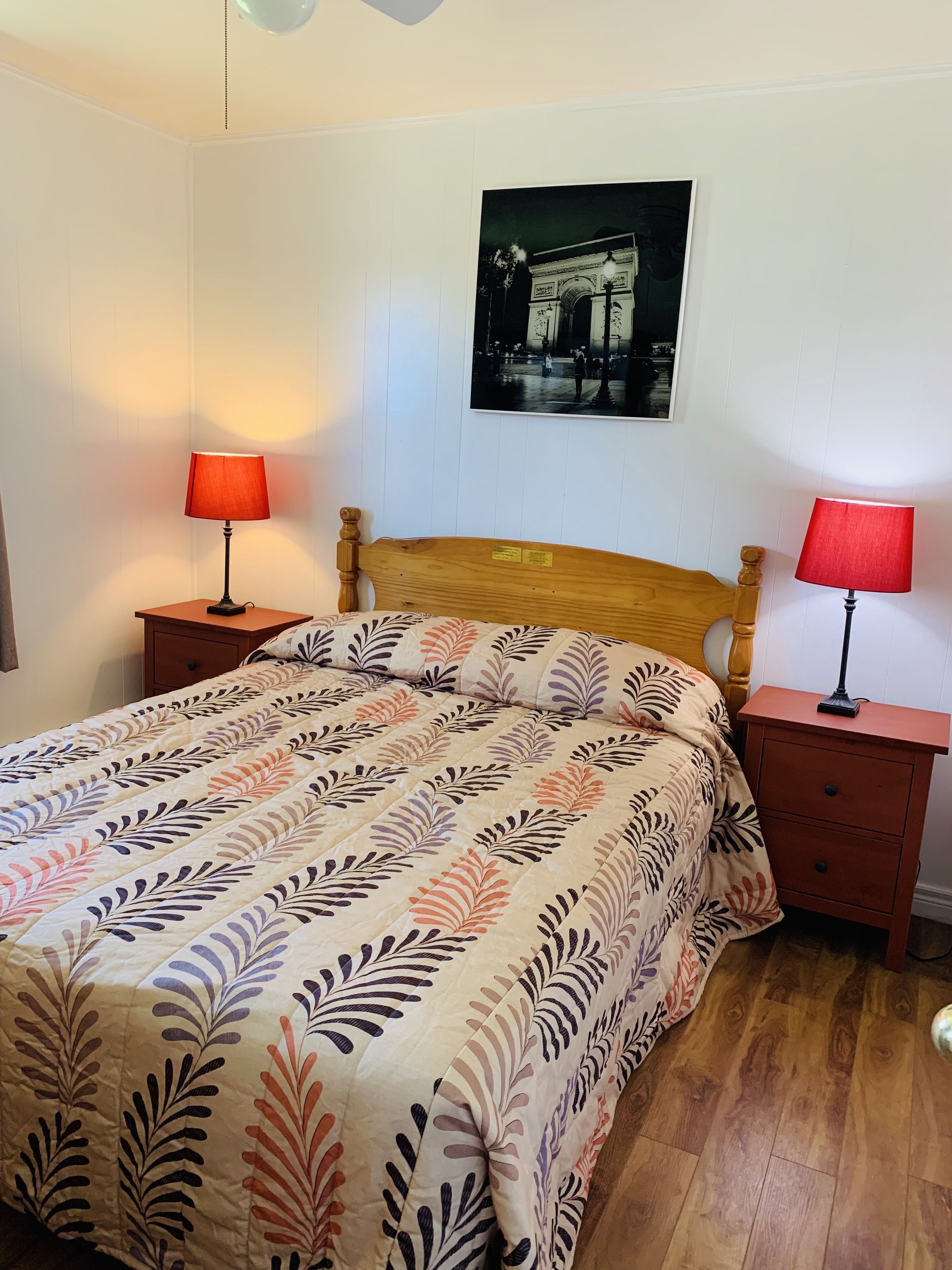 A bedroom with a wooden bed, two bedside tables with lamps, a wall-mounted picture, and a wooden floor.