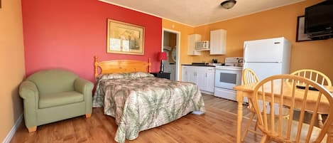 A room with a bed, a green armchair, a wooden dining table with chairs, a kitchen area with a refrigerator, stove, and microwave.