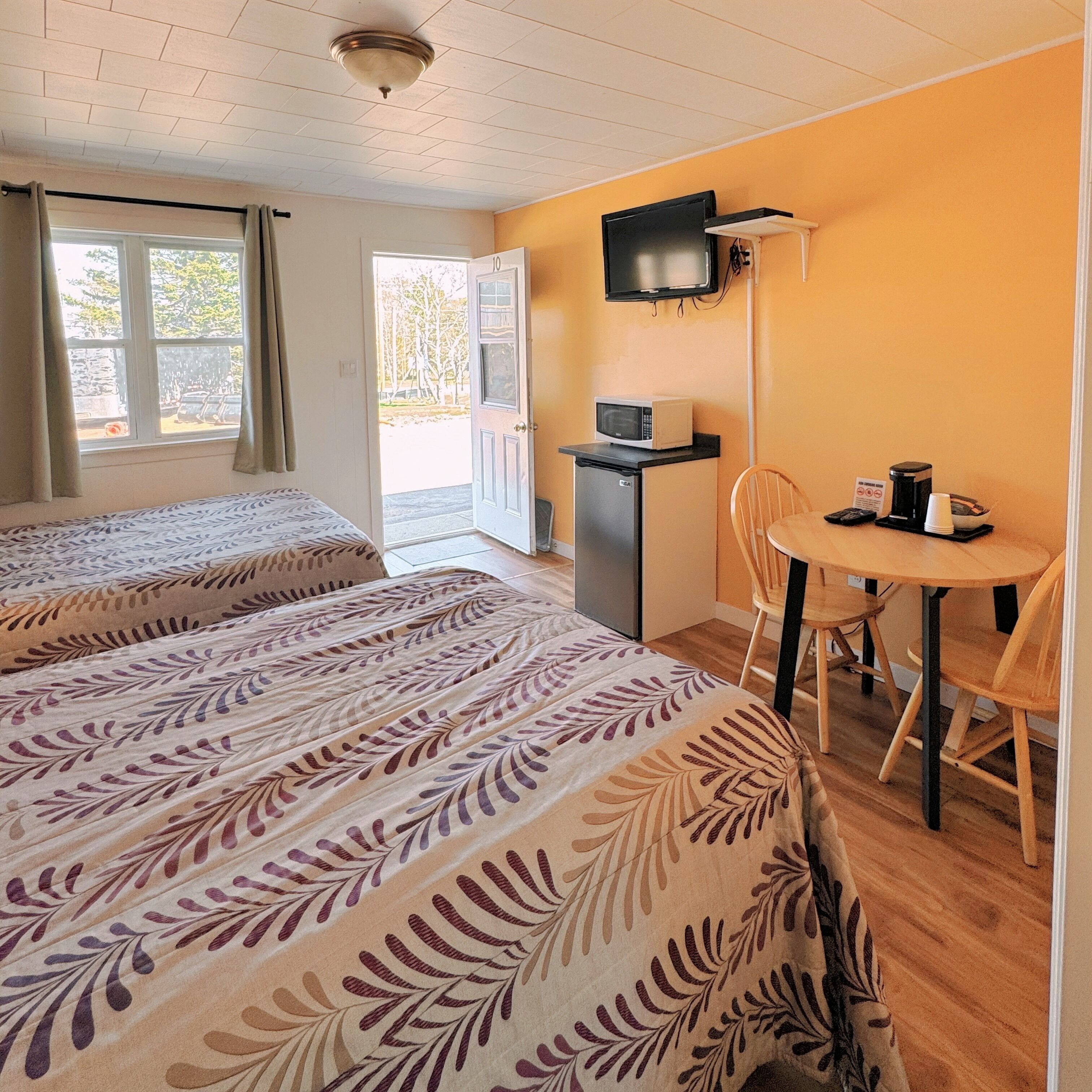Quarto de hotel com duas camas, uma kitchenette pequena, uma televisão e área de refeições com mesa e cadeiras.