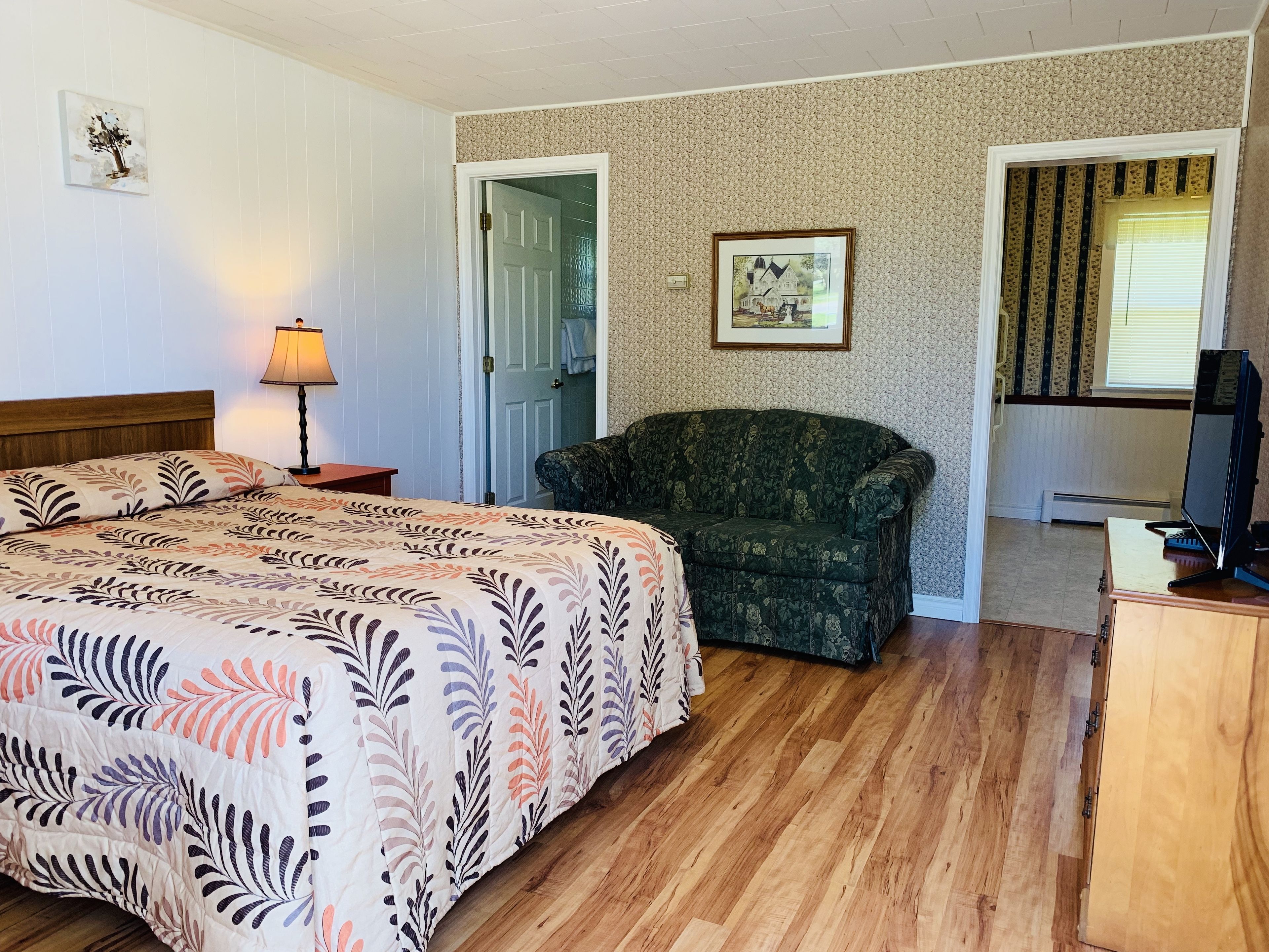 A hotel room with a bed, a chair, a desk with a computer, a lamp, and a framed picture on the wall.