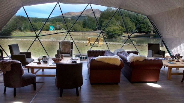 Amplio interior con un gran ventanal que ofrece vistas a un lago y colinas boscosas. Amueblado con sofás y sillones de cuero, la habitación incluye una mesa de madera con varios objetos.